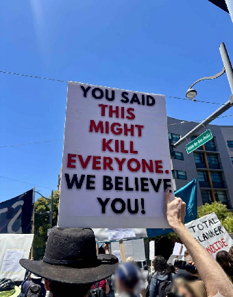 A protester outside OpenAI's San Francisco office holds a hand-lettered sign reading: 'You said this might kill everyone. We believe you!'